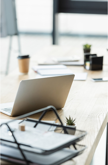 An open laptop sits on a bright wooden desk with documents, a coffee cup, small potted plants, and office supplies, reflecting a modern workspace where Our Services help keep you organized and productive.