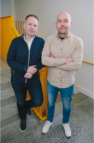 Two men stand on an indoor staircase with yellow railings, embodying confidence and ease. One wears a navy jacket, the other a beige shirt with arms crossed. Both look at the camera, reflecting a modern vibe suited for innovative digital experiences.